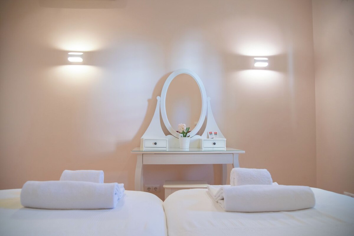 A hotel room with two beds, a white vanity with a mirror, and two wall-mounted lights.