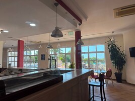 Uma área de bar moderna com um balcão amplo, iluminação pendente e vista para o exterior através de grandes janelas.