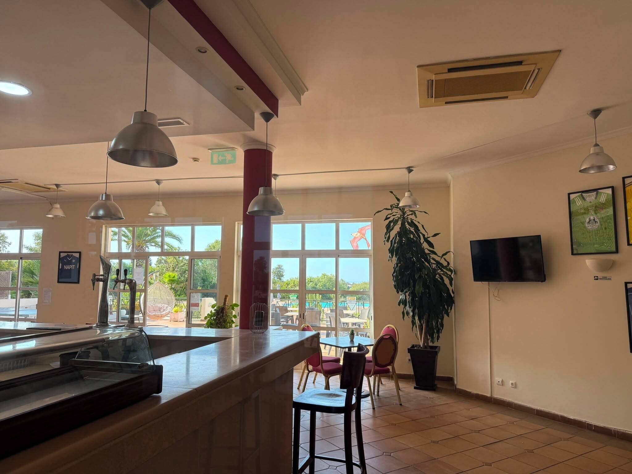 A restaurant with a counter, chairs, hanging lights, a TV, and large windows with a view.
