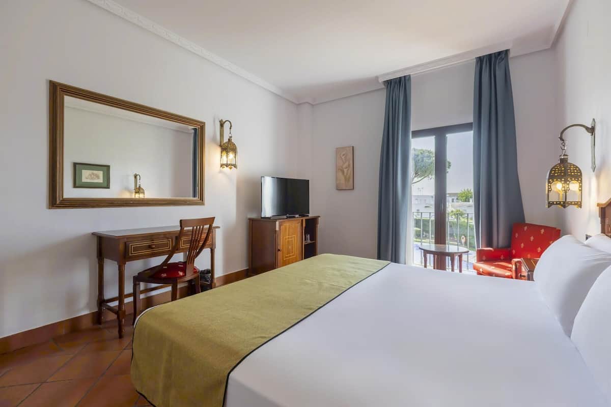 A hotel room with a large bed, a desk with a television, a chair, a small table, and a balcony with curtains.