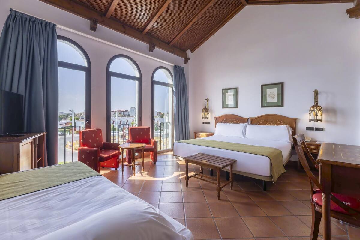 A hotel room with a large bed, a smaller bed, a desk, a chair, a table, and a balcony with a view of the city.