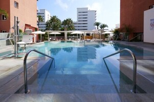Een zwembad met helder water, omringd door ligbedden en parasols, met gebouwen op de achtergrond.