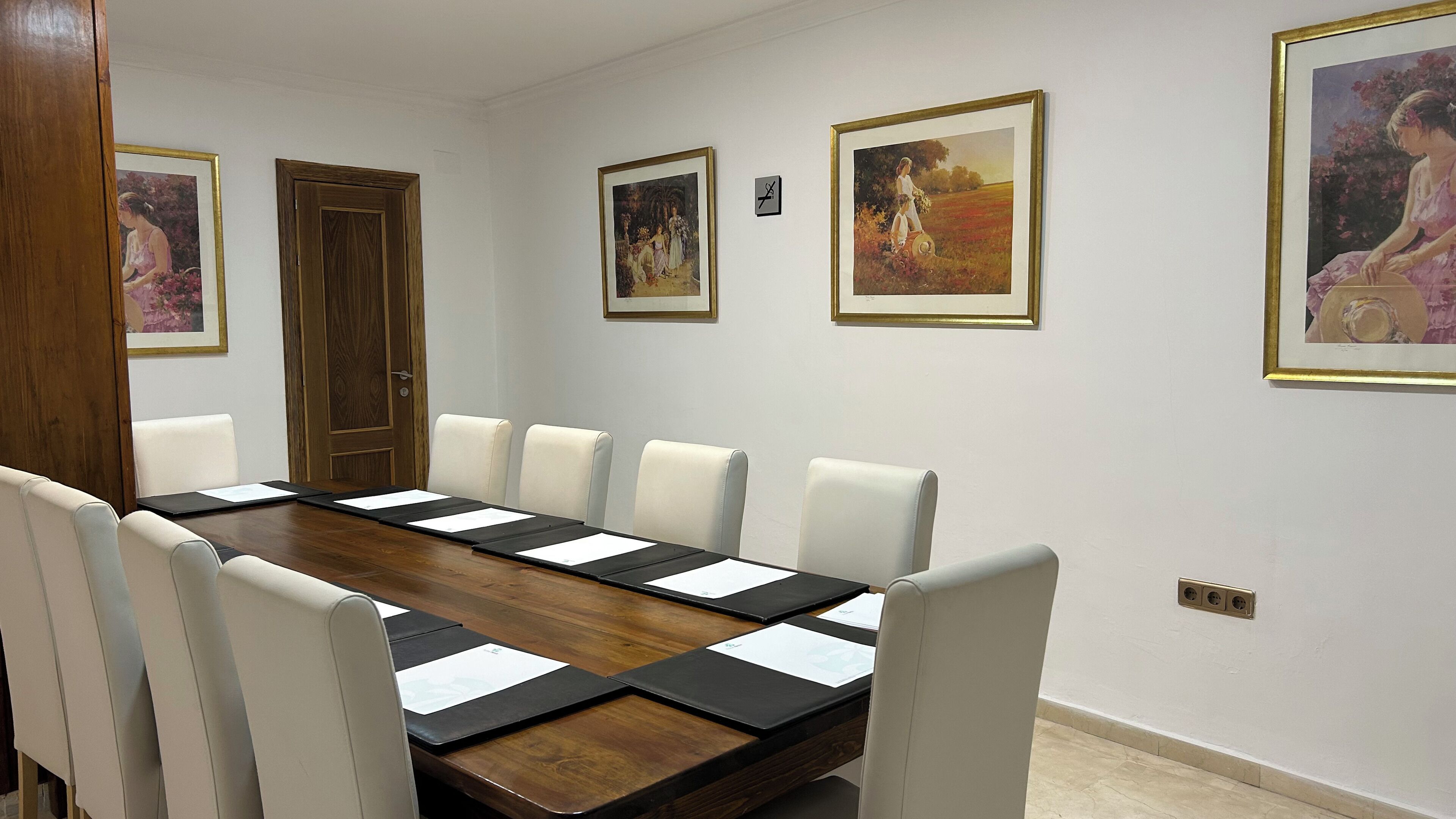 A conference room with a long wooden table, six white chairs, and framed artworks on the walls.