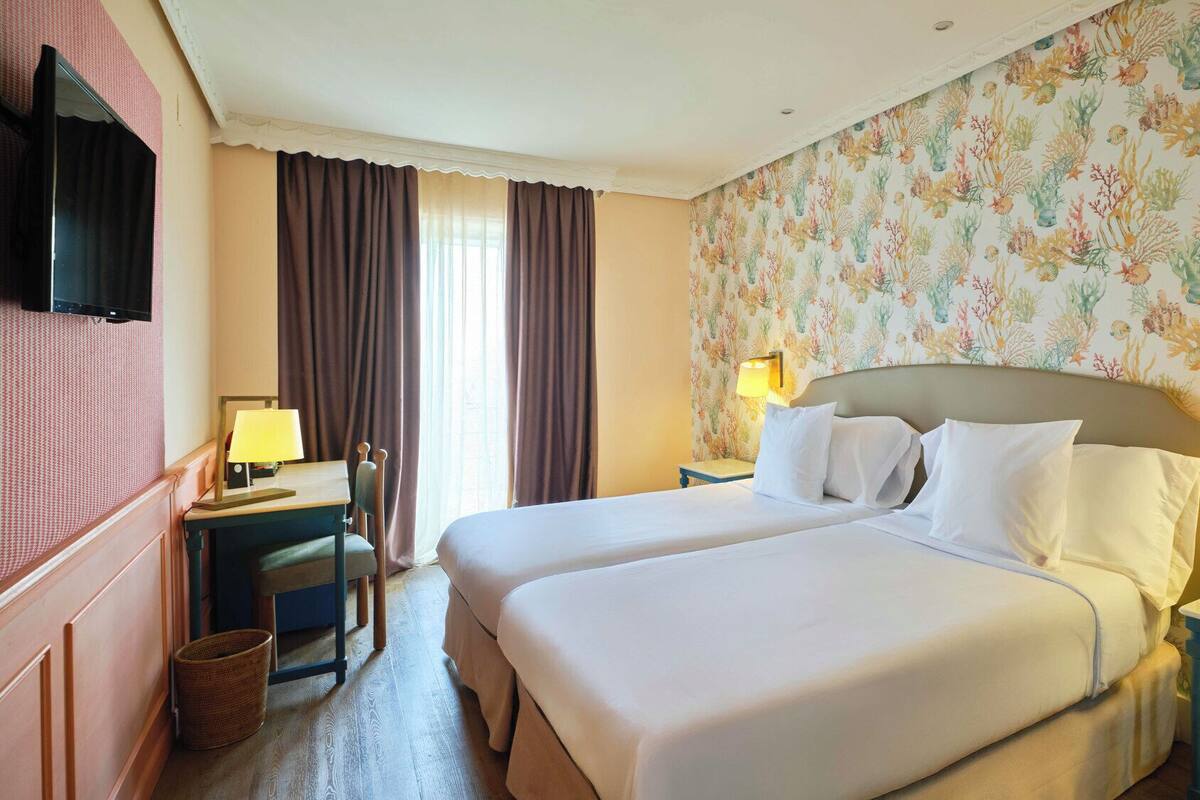 Ein Hotelzimmer mit zwei Betten, einem Schreibtisch, einem Sessel, einem Fernseher und einer Wand mit floralem Muster.
