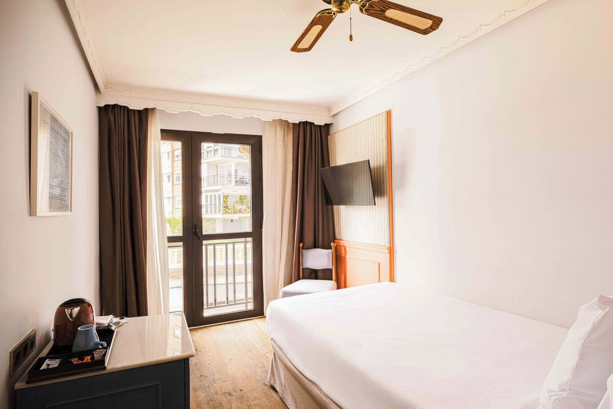 Ein Hotelzimmer mit Bett, Schreibtisch mit Wasserkocher, Flachbildfernseher und Balkon mit Vorhängen.