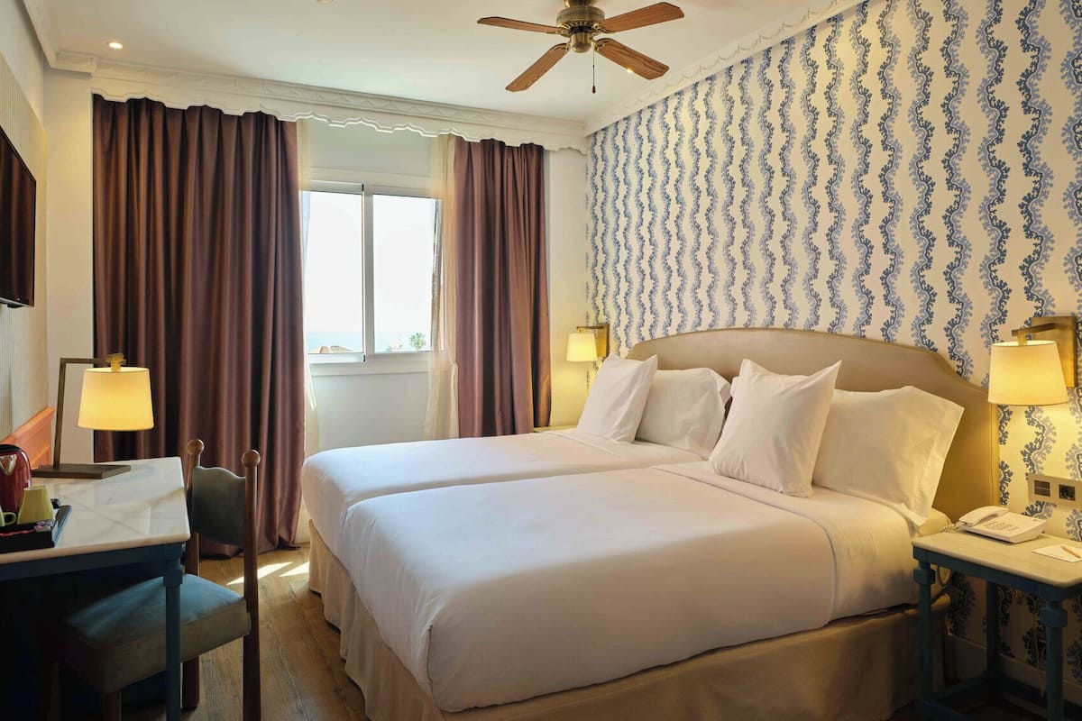 Ein Hotelzimmer mit einem Doppelbett, einem Schreibtisch mit Lampe, einem Sessel, einem Telefon und einem Deckenventilator.