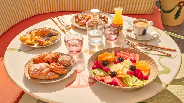 Une table ronde, dressée à l’extérieur avec une vue sur la mer, sur laquelle sont disposés divers aliments, notamment des pâtisseries et des fruits.