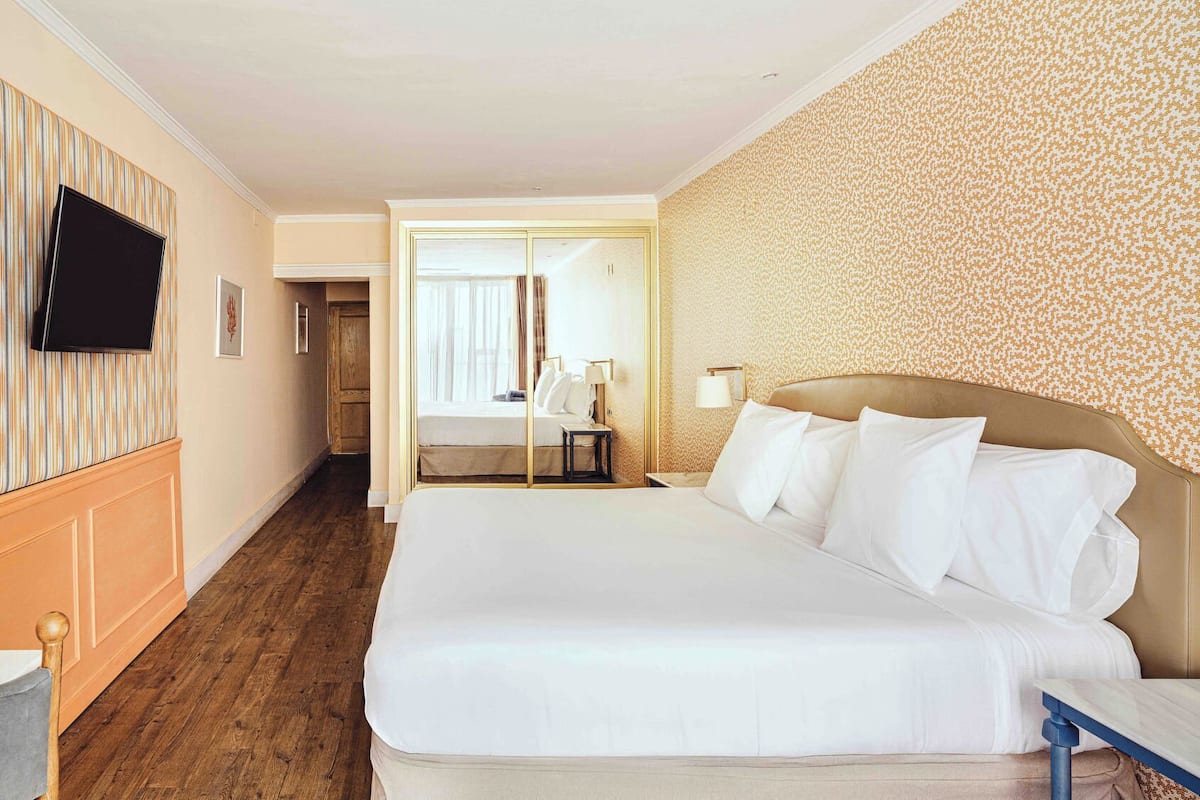 Ein Hotelzimmer mit einem großen Bett, einem an der Wand montierten Fernseher, Holzboden und einer Wand mit ornamentalem Wandpapier.