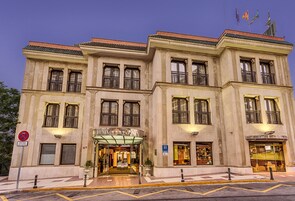 Een groot, historisch gebouw met meerdere balkons, een centrale ingang met glazen overkapping en een bord dat een hotel aangeeft.