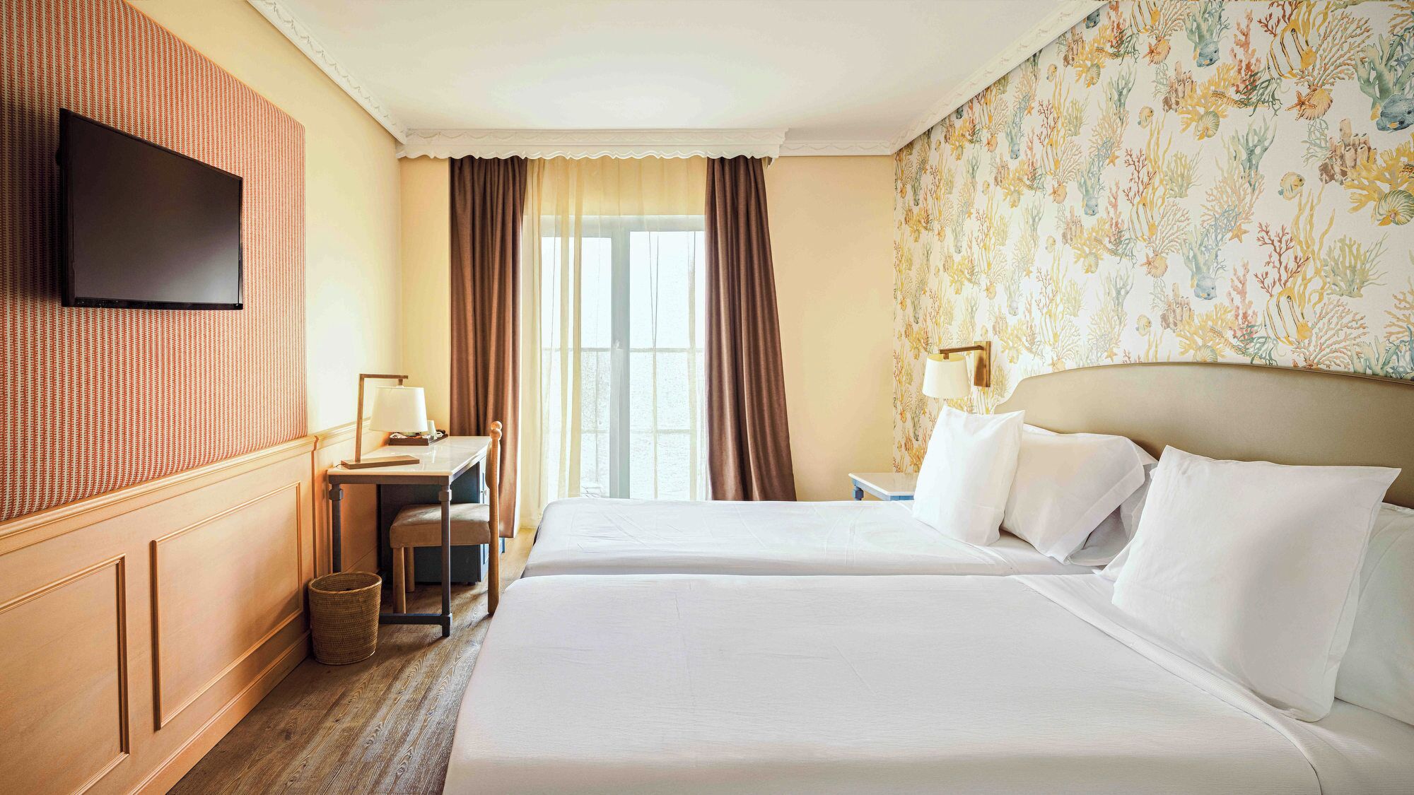 A hotel room with two beds, a desk, a chair, a television, and a window with curtains.
