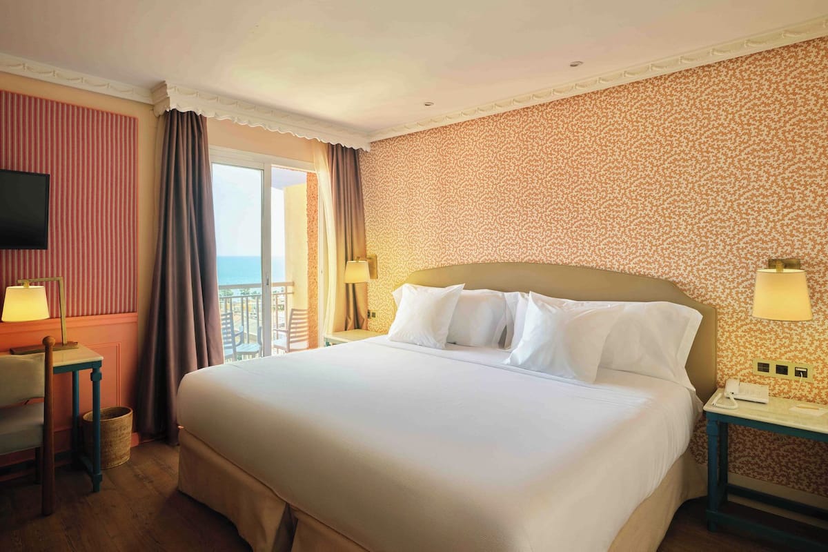 Ein Hotelzimmer mit einem großen Bett, einem Schreibtisch, einem Stuhl, einem Fernseher und einem Balkon mit Vorhängen.