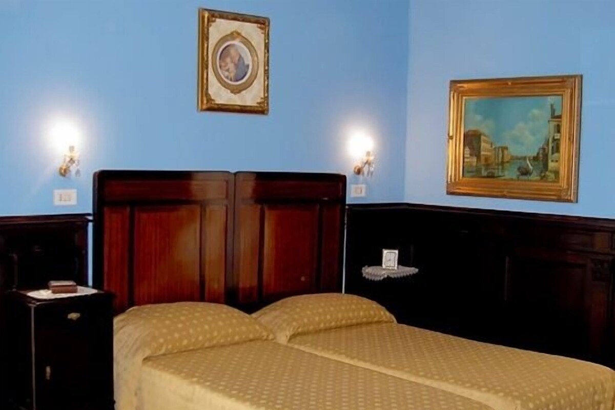 A room with two beds, wooden headboards, a nightstand, a framed picture, and wall-mounted lights.