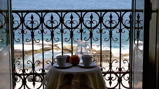 A balcony with a table set for two, overlooking a coastal view with a sailboat on the water. - Hotel Aurora (Syracuse)