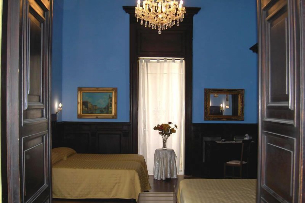 A room with two beds, a chandelier, a painting, a mirror, and a vase with flowers.