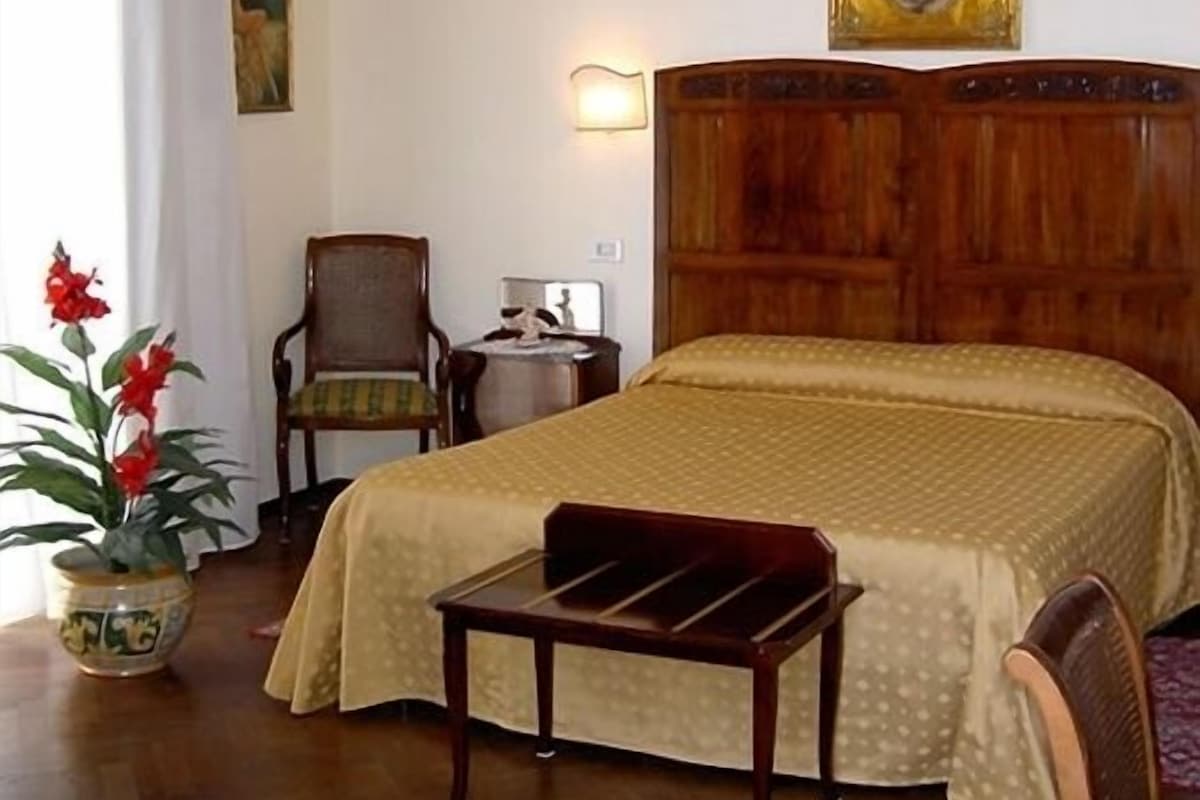 A hotel room with a wooden bed, a chair, a small table, and a potted plant.