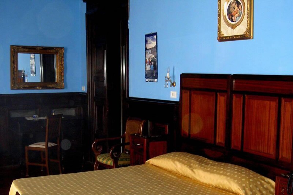 A hotel room with a large bed, a wooden headboard, a desk with a chair, a mirror, and a framed picture on the wall.