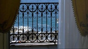 A view through a window with curtains, revealing a balcony with a metal railing and the sea in the background. - Hotel Aurora (Syracuse)
