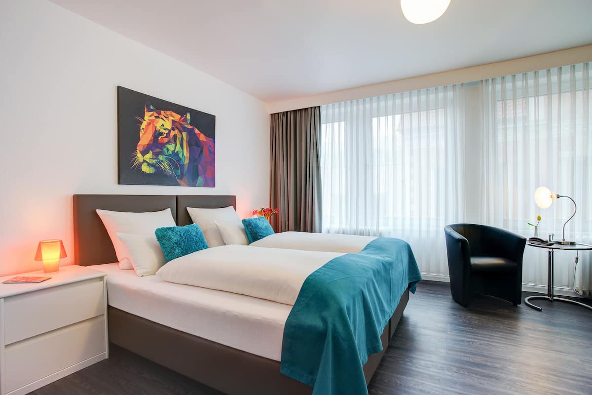 A hotel room with a bed, a nightstand, a chair, a table, and a painting on the wall.