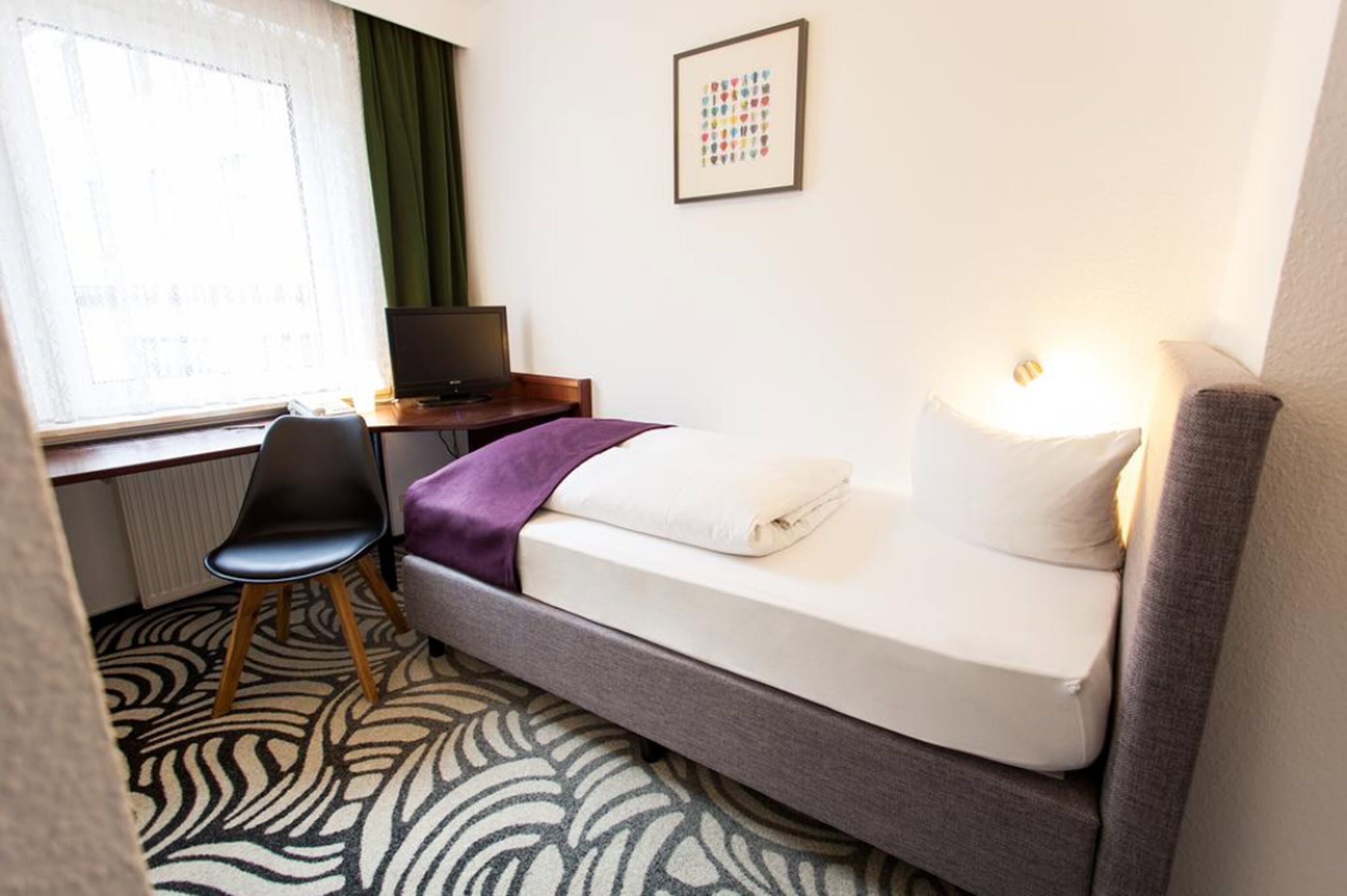 A hotel room with a bed, a desk with a computer, a chair, and a window with curtains.