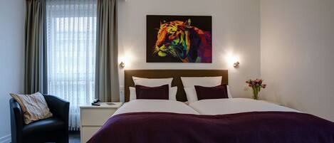 A hotel room with a large bed, a nightstand, a chair, a window with curtains, and a wall-mounted painting.
