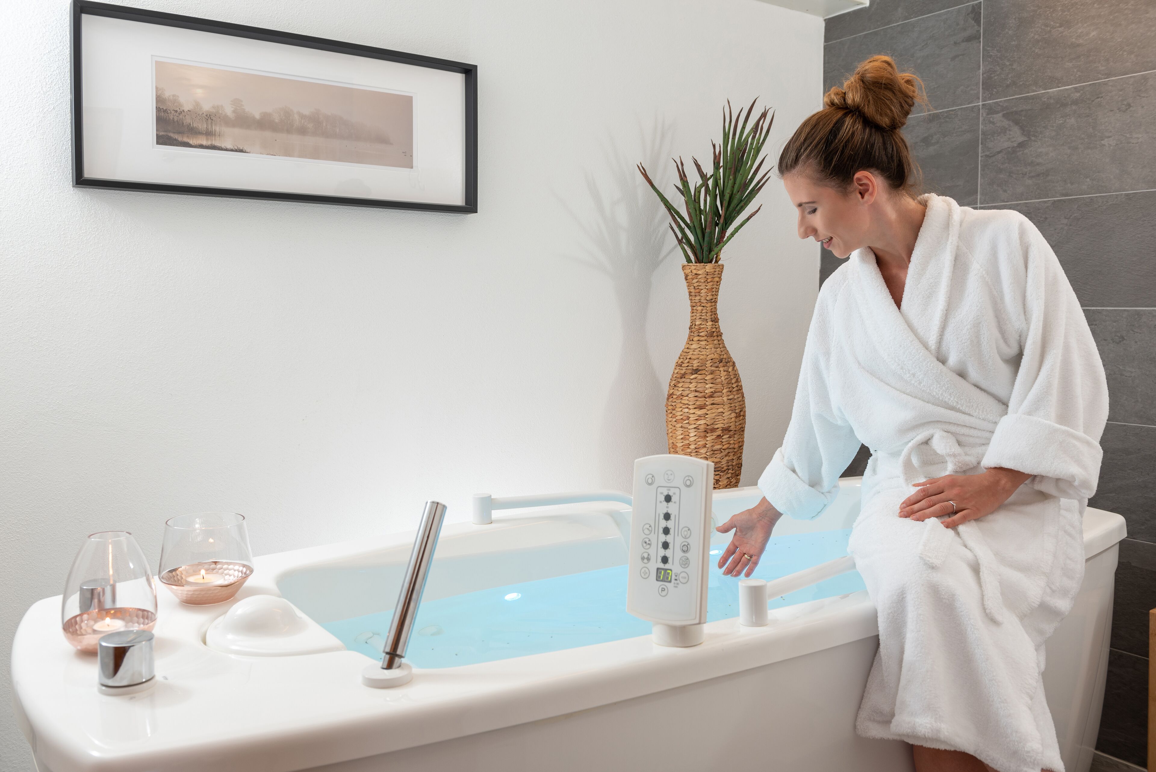A person in a white bathrobe sitting in a bathtub with a control panel, a framed picture on the wall, and a plant beside the tub.