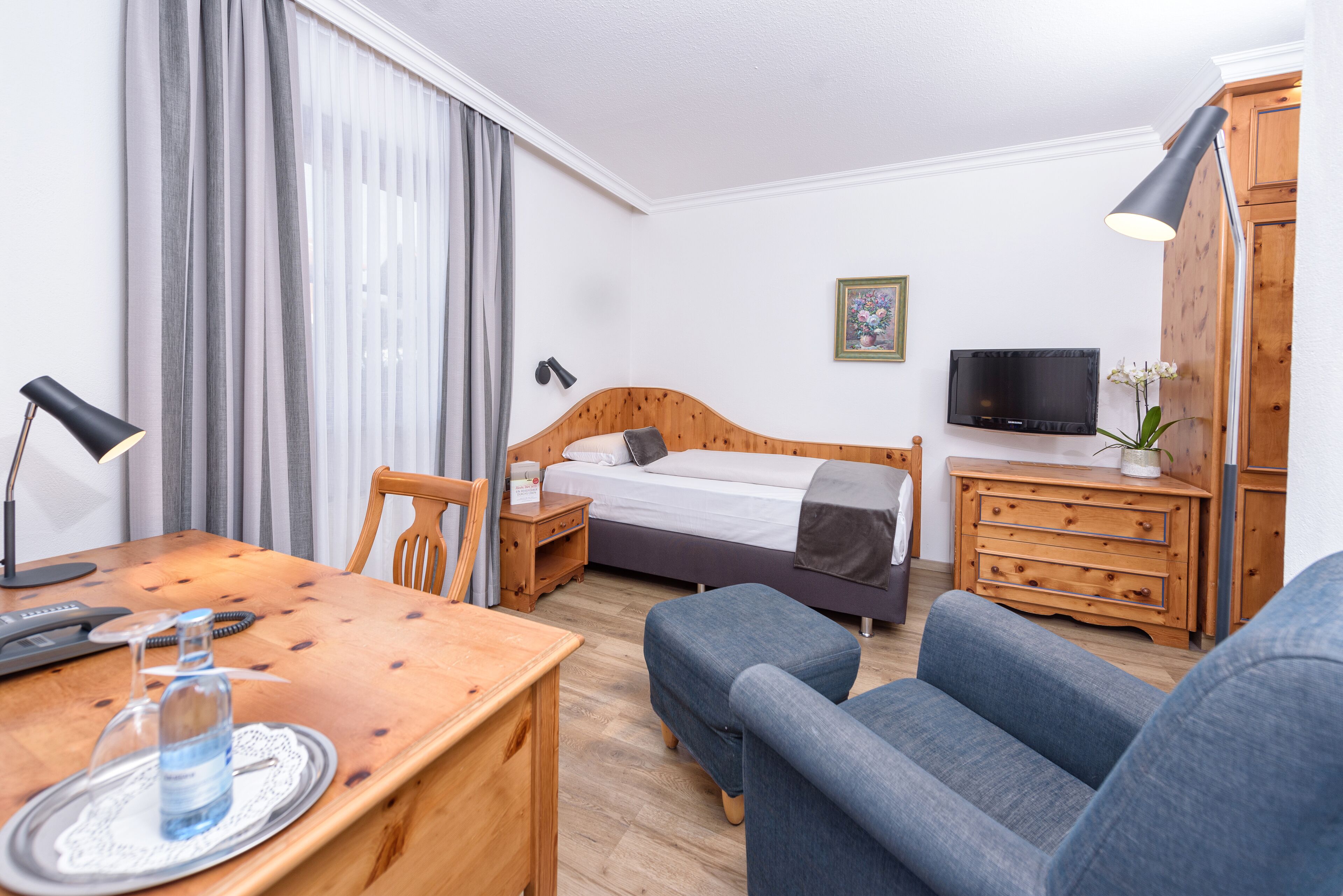 Habitació d'hotel amb un llit de fusta, un escriptori amb una làmpada, una cadira, un televisor i un armari.