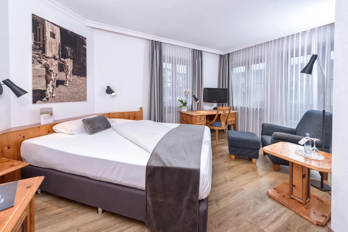A hotel room with a bed, a desk with a computer, a chair, a TV, and a large window with curtains.