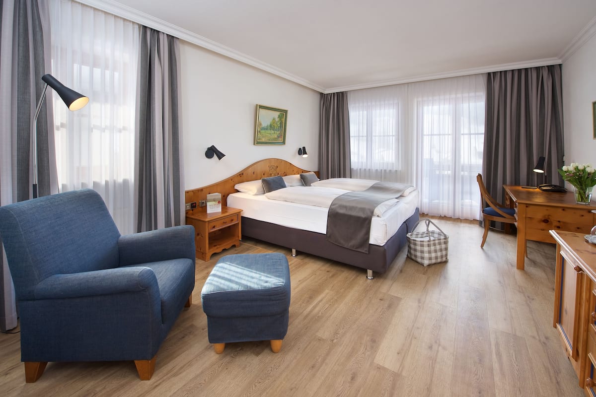 A hotel room with a bed, a blue armchair, a wooden desk, and a small ottoman.