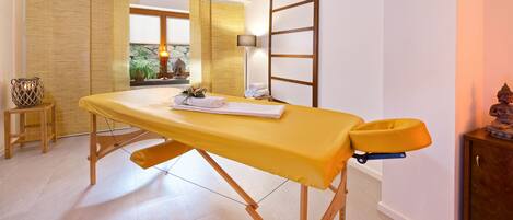 A room with a massage table, a small wooden cabinet, a lamp, and a window with a view of greenery.