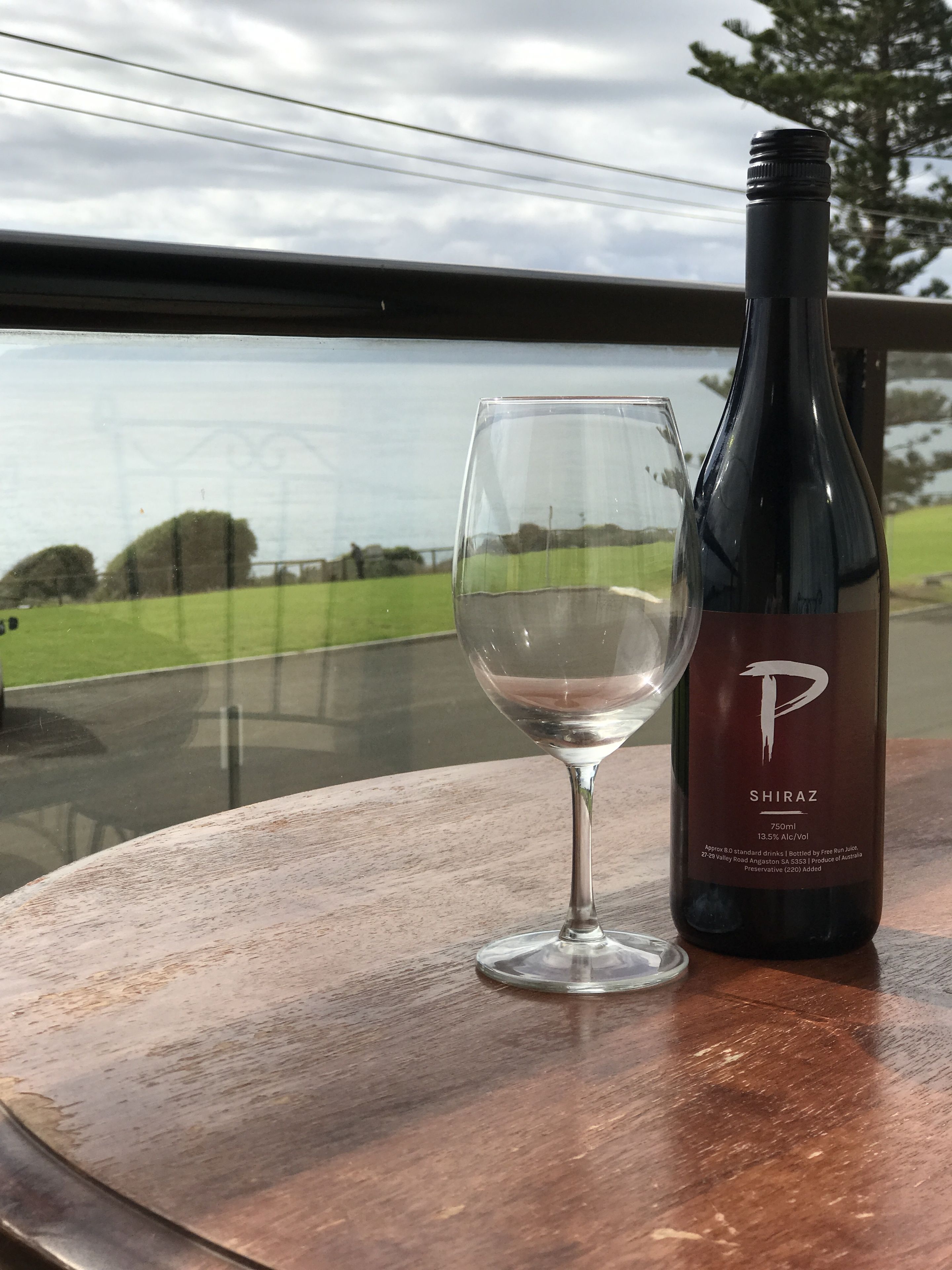 Une bouteille de vin Shiraz et un verre vide sur une table, avec vue sur un paysage verdoyant par la fenêtre.