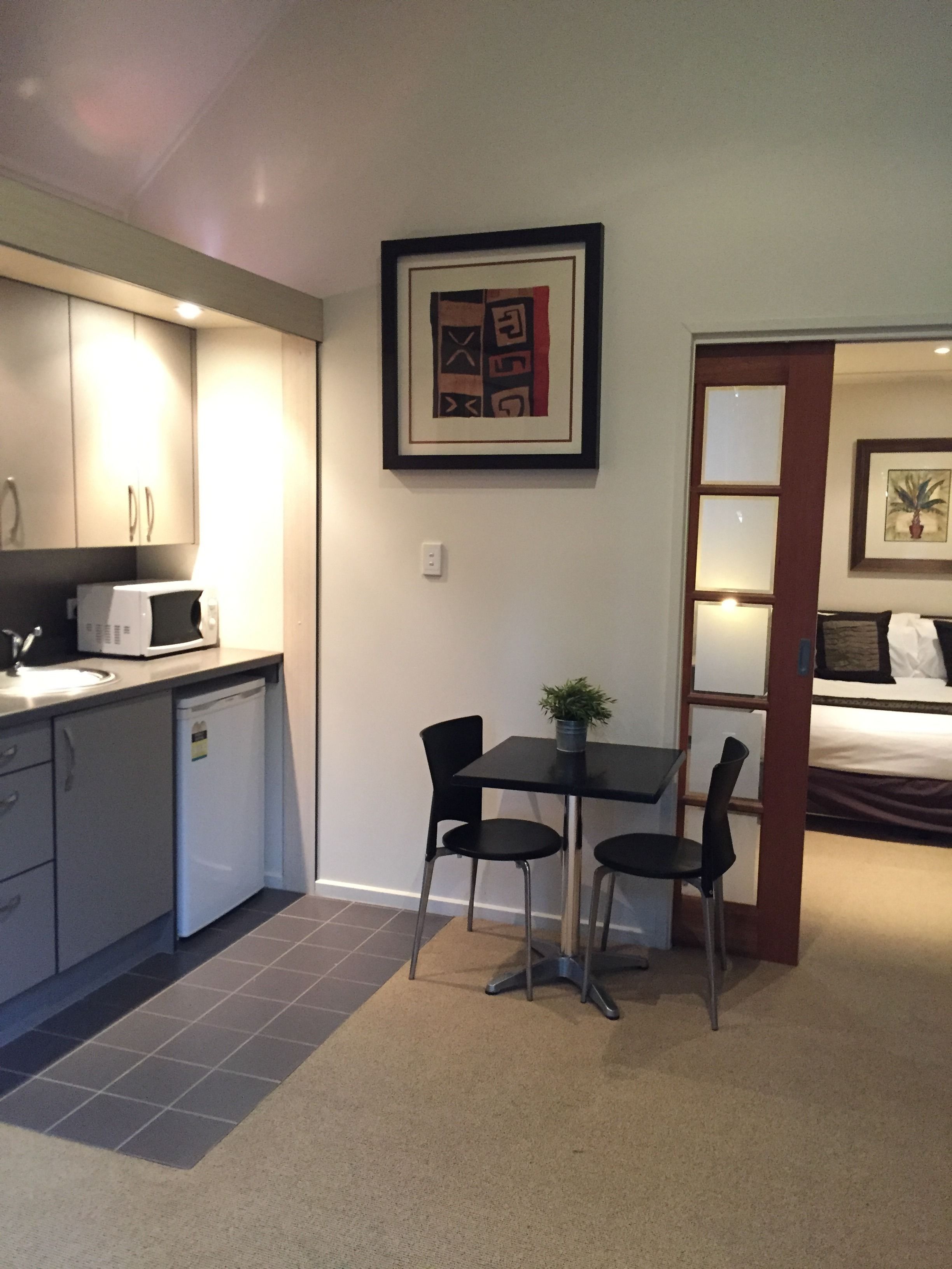 A compact kitchen with a microwave, sink, and cabinets. There is a dining area with a table and chairs, and a small potted plant on the table. A framed artwork is on the wall above the dining area.