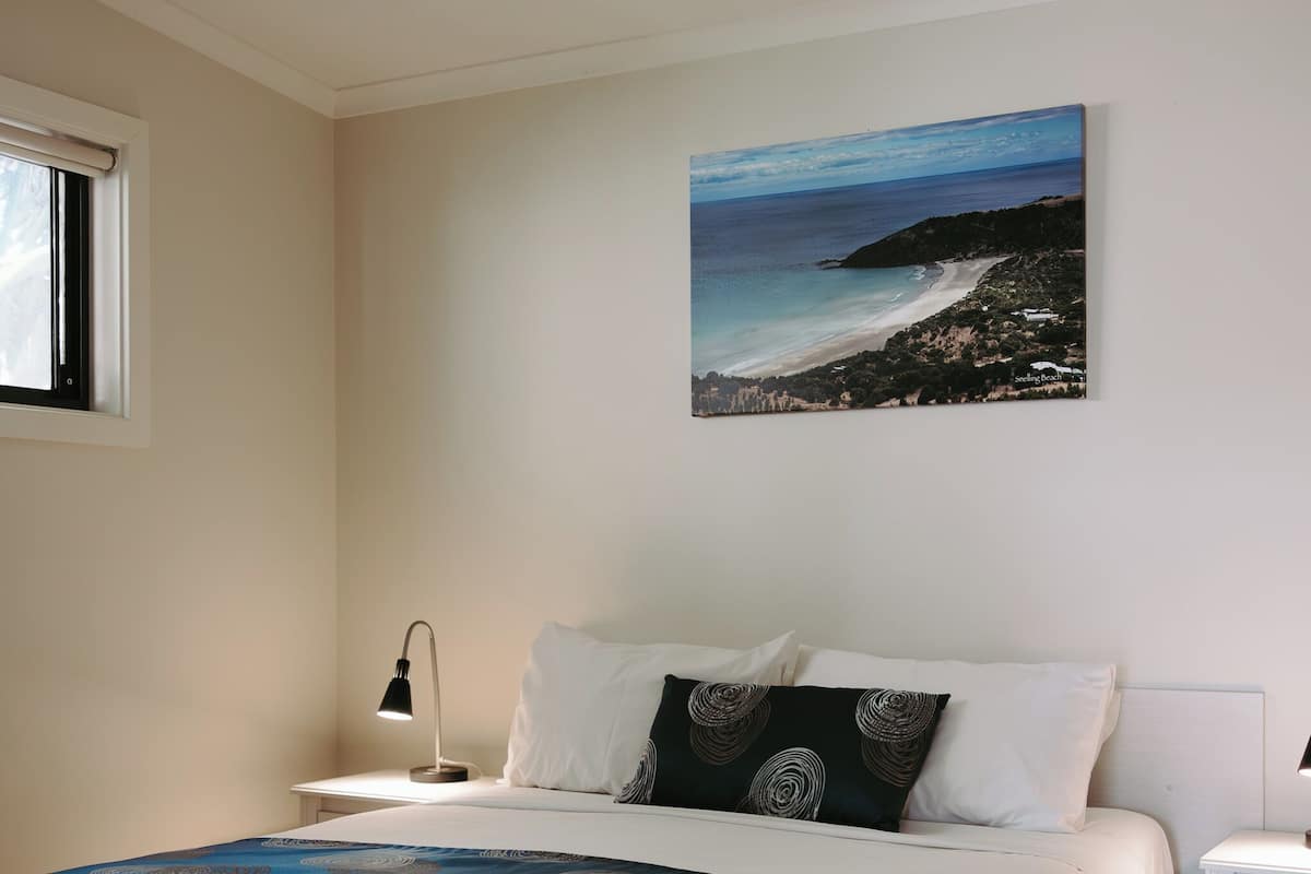 Une chambre à coucher comprenant un lit, une table de chevet, une lampe fixée au mur, un ventilateur de plafond et un tableau représentant une vue sur la plage.