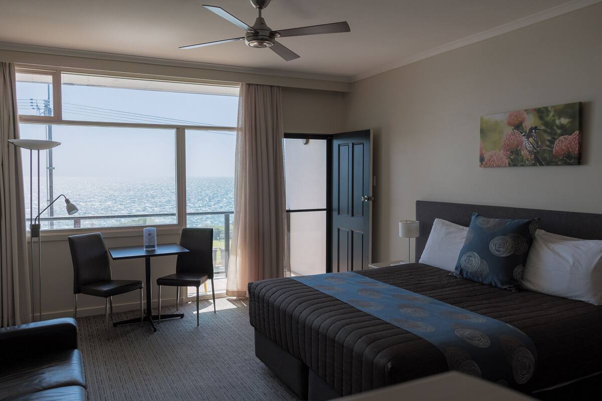 Une chambre d’hôtel comprenant un lit, un bureau, deux chaises, un ventilateur de plafond, une grande fenêtre donnant sur la mer et un tableau au mur.