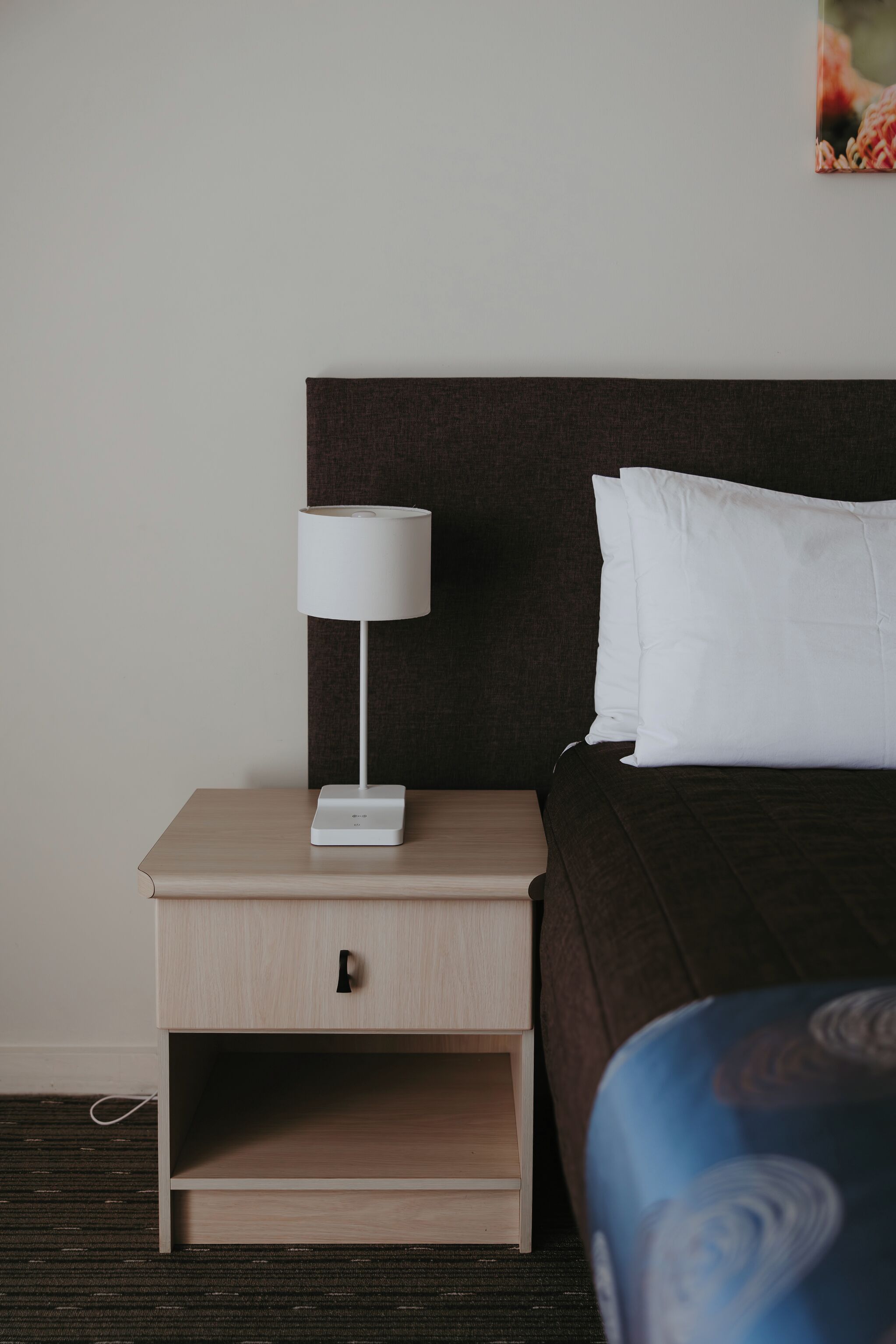 Una cama bien hecha con almohadas blancas y un cabecero oscuro, una mesita de noche pequeña con una lámpara blanca y un pequeño mueble auxiliar de madera clara.