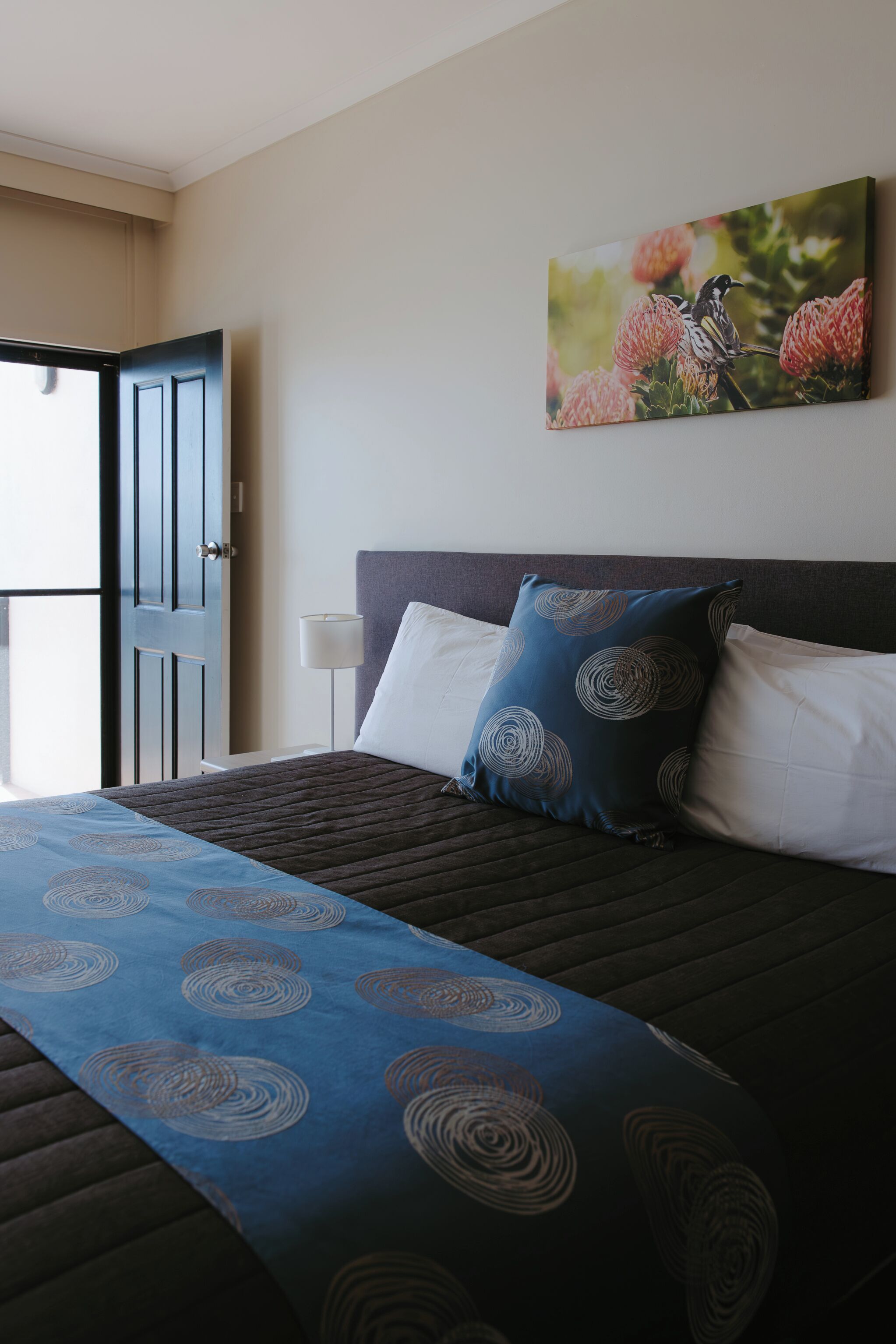 Habitación de hotel con una cama, un cuadro de flores, una mesita de noche y una puerta.