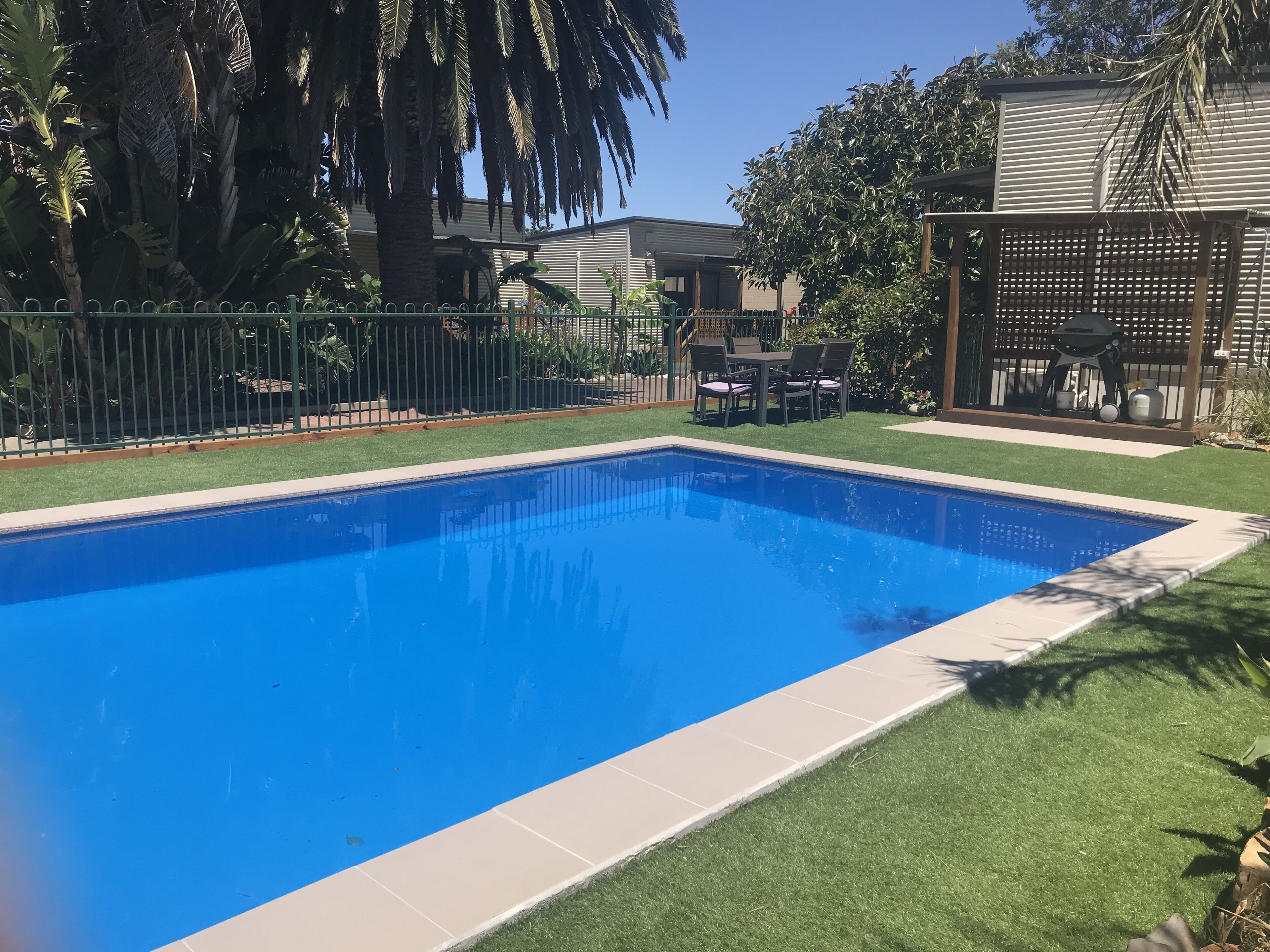 A rectangular swimming pool with a tiled edge, surrounded by artificial grass and a metal fence.