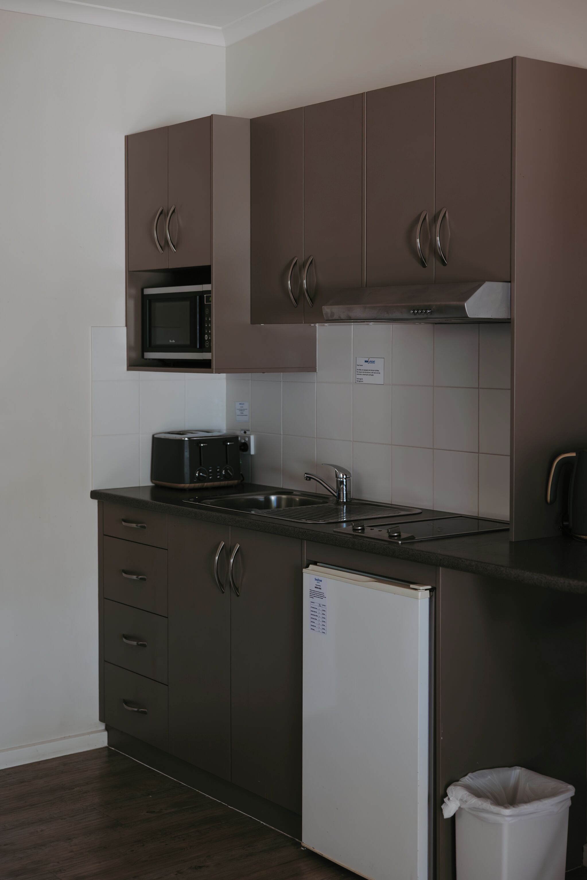 Une cuisine de taille réduite, dotée de meubles de rangement sombres, d’un four à micro-ondes, d’un grille-pain, d’un évier et d’un réfrigérateur.