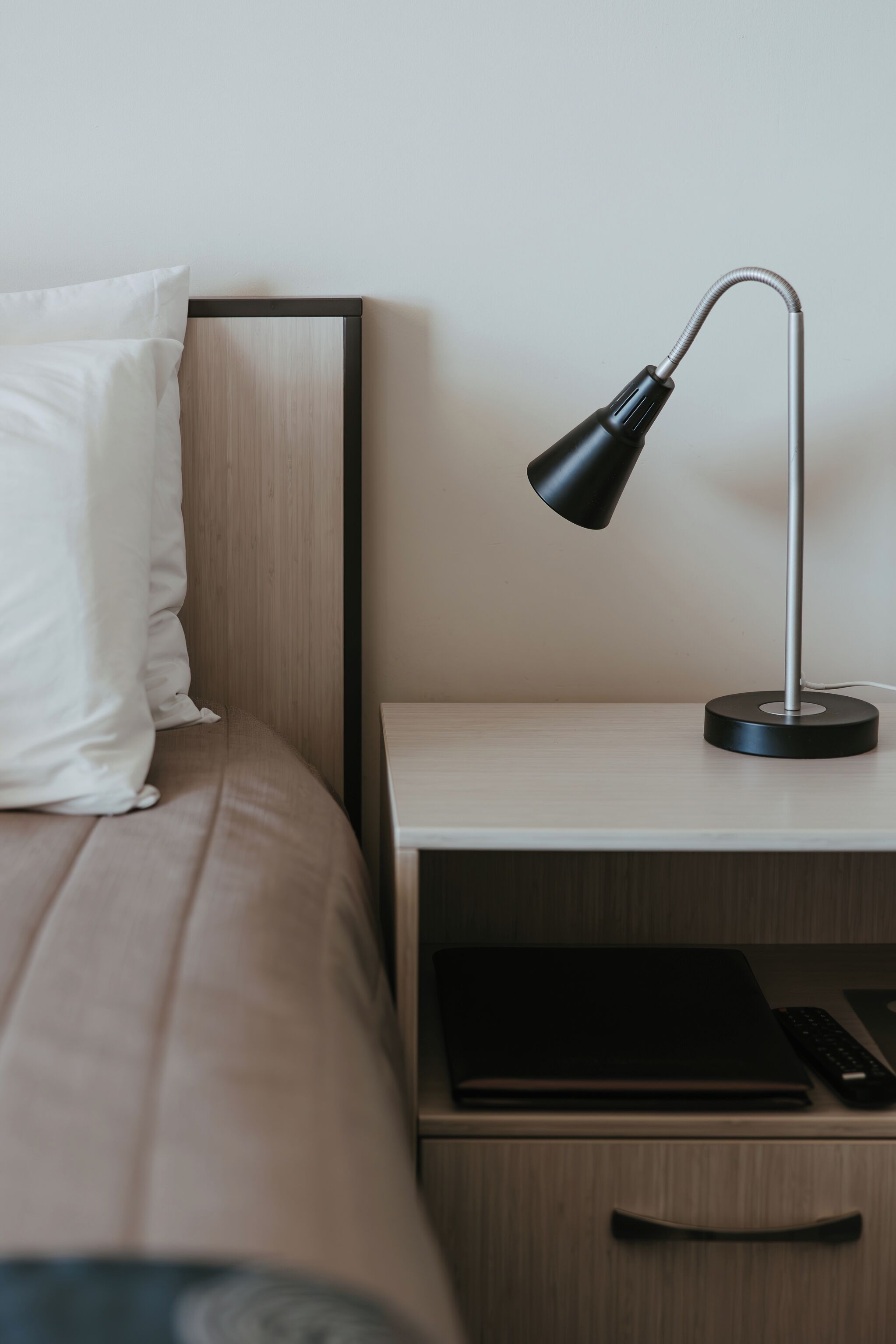 A neatly made bed with a white pillow, a bedside table with a black lamp, and a drawer with a black object on it.