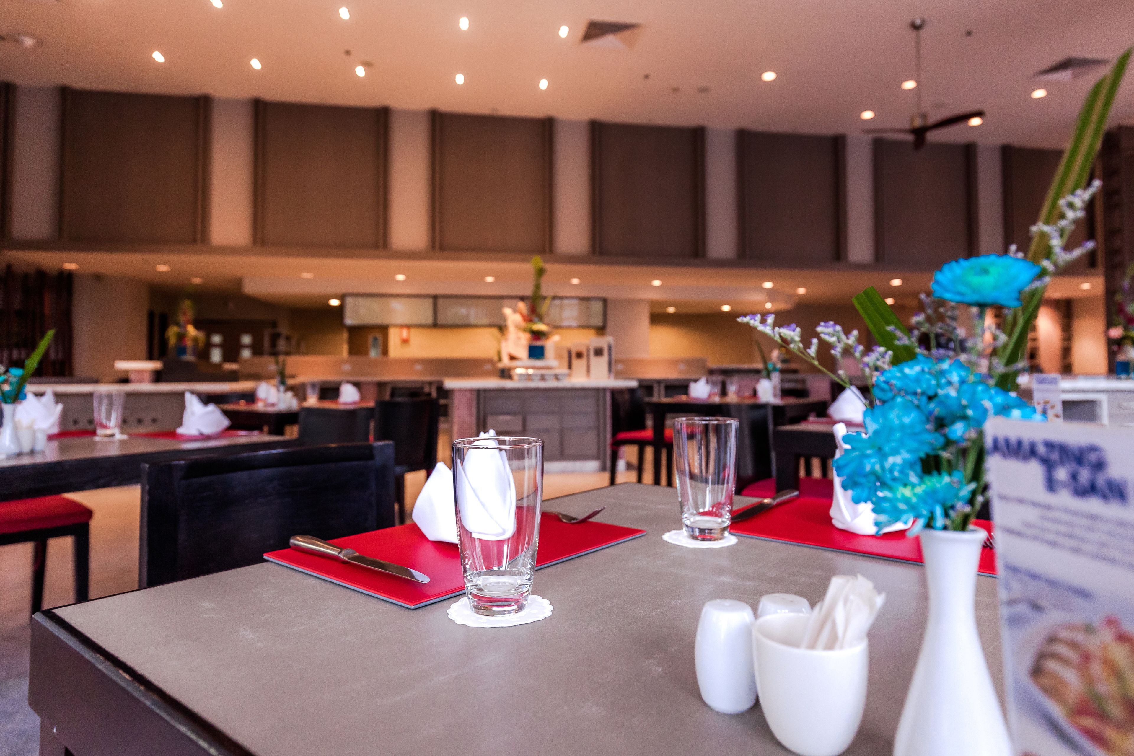 A restaurant with tables set with red placemats, clear glasses, and a vase with blue flowers.