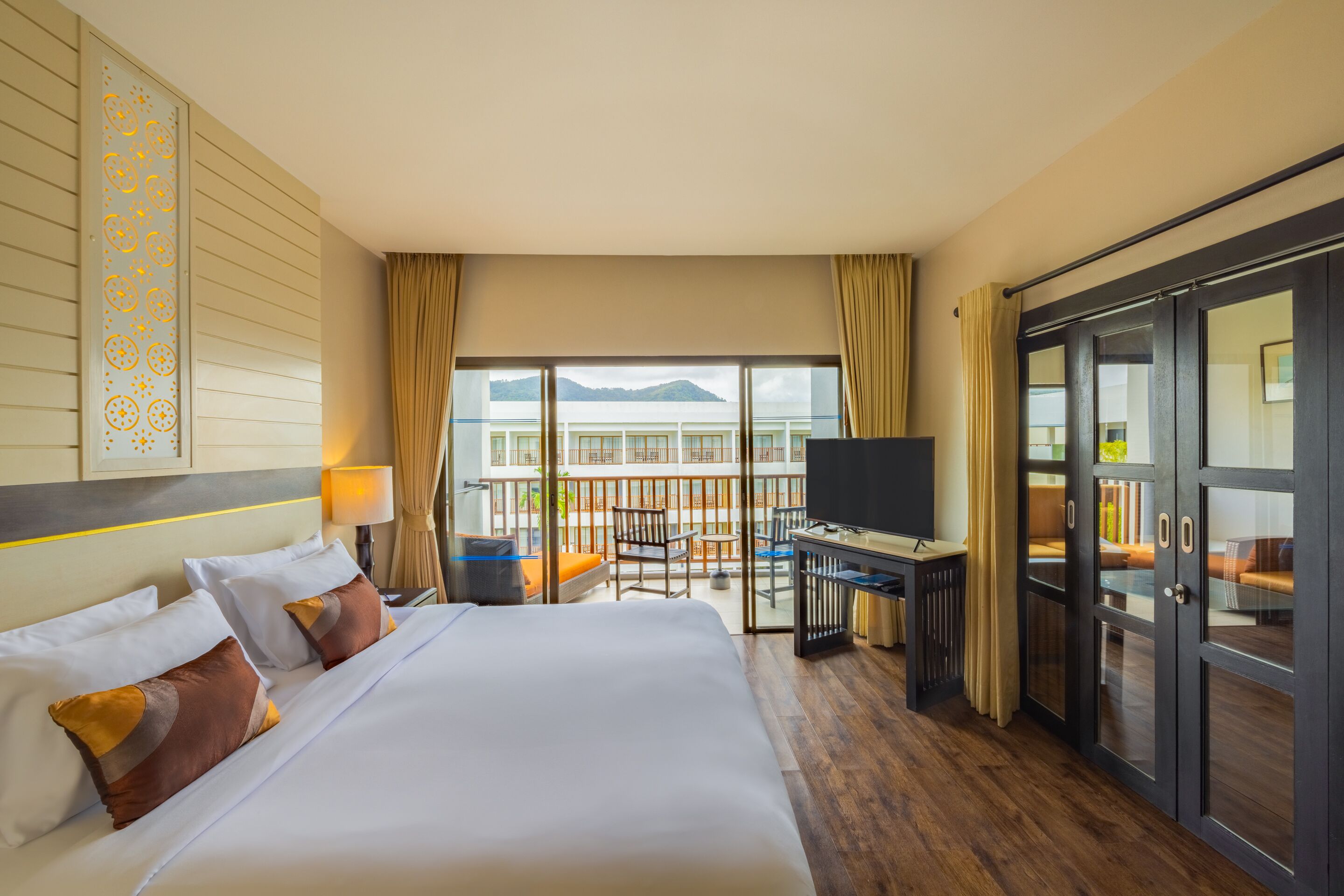 A hotel room with a large bed, a desk, a TV, and a balcony with a view of the sea.