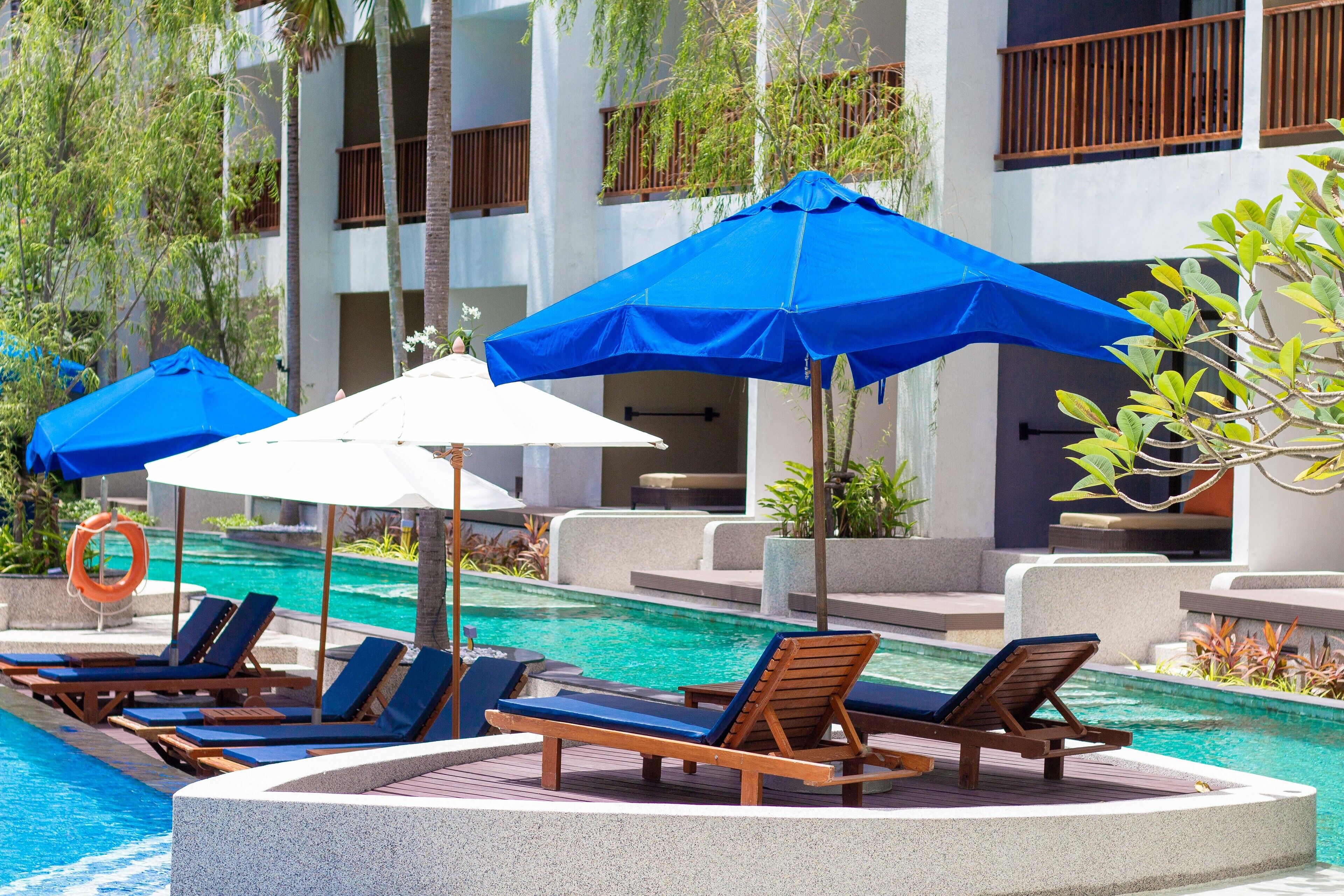 A poolside area with lounge chairs and umbrellas, surrounded by a building with balconies and plants.