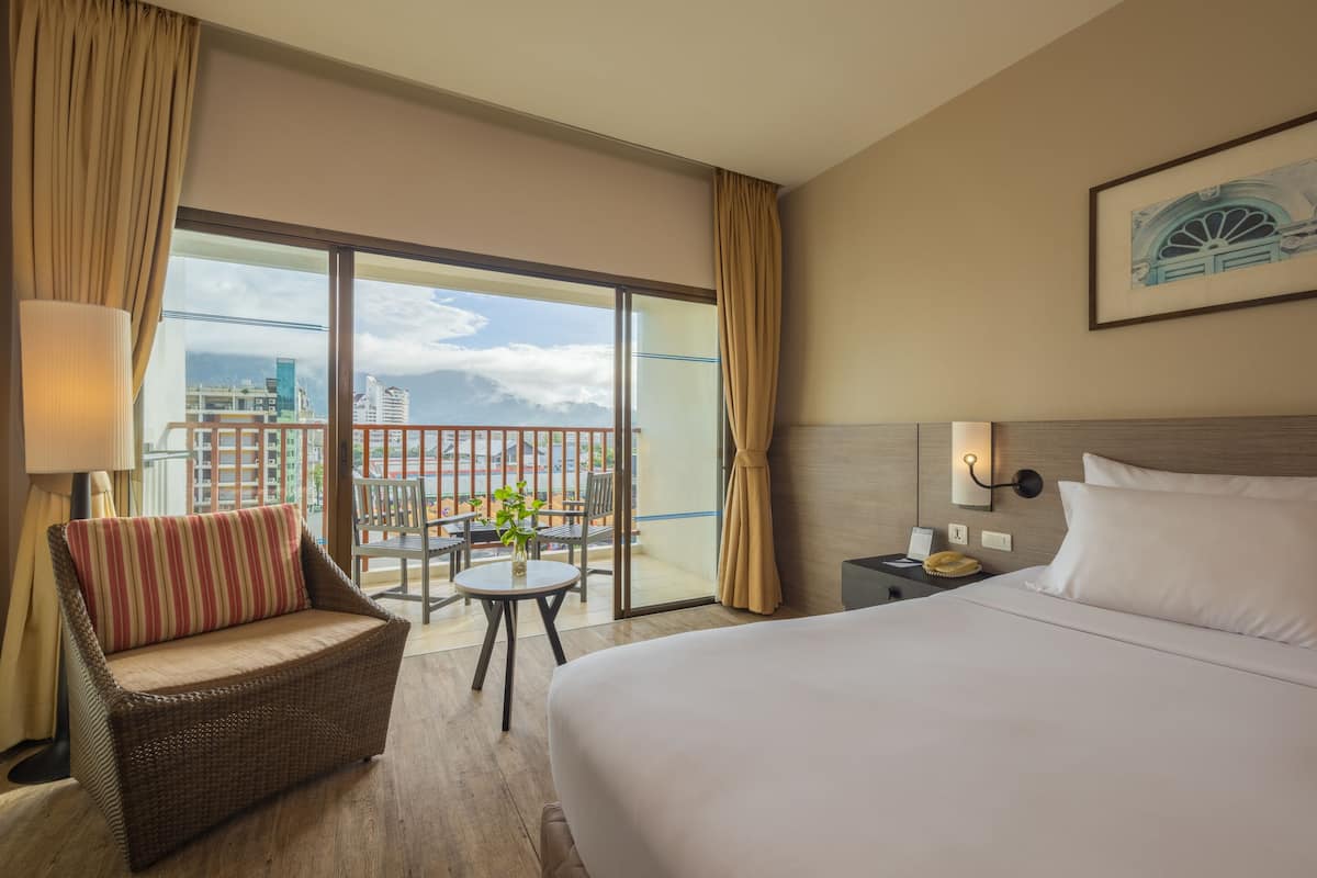 Hotellihuone, jossa on suuri sänky, sohva, pieni pöytä, jossa on kasvi, yöpöytä ja parvekkeelta avautuva näkymä kaupunkiin.