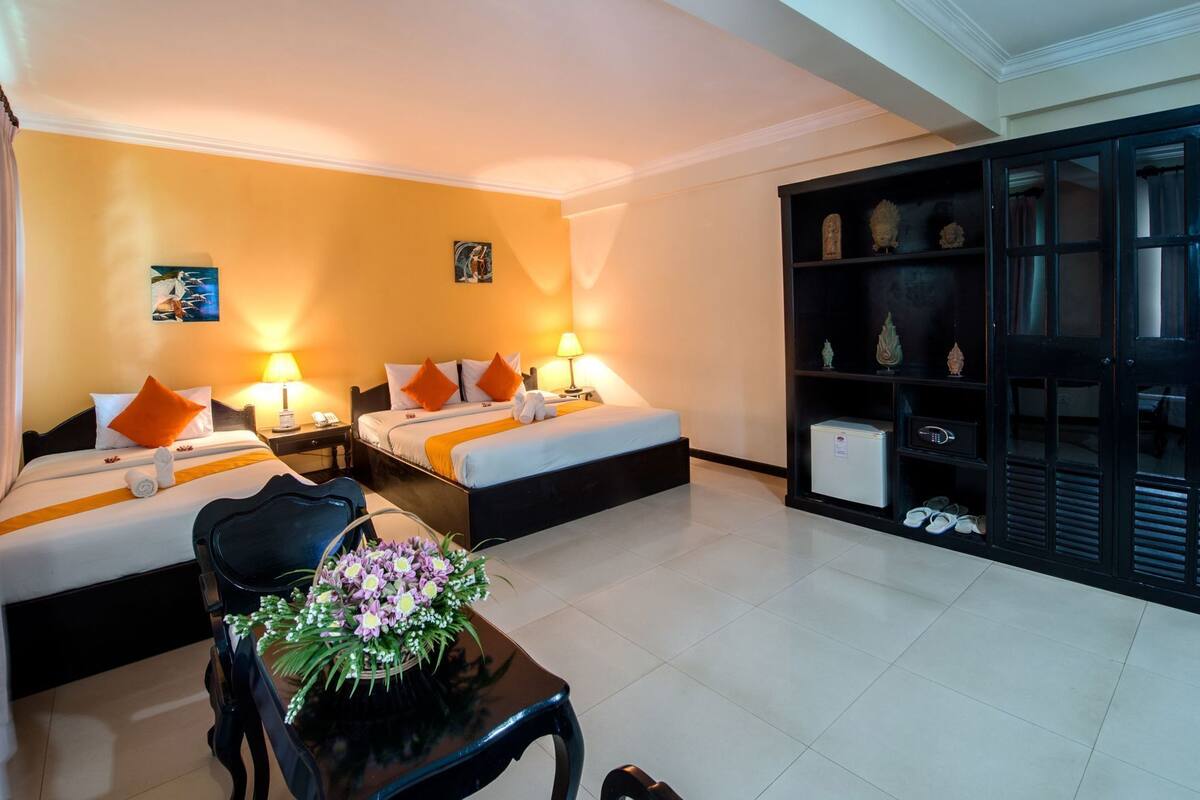 Ein Hotelzimmer mit zwei Betten, einem Schreibtisch, einem Stuhl, einem kleinen Tisch mit Blumen und einer schwarzen Kommode mit Dekoration.