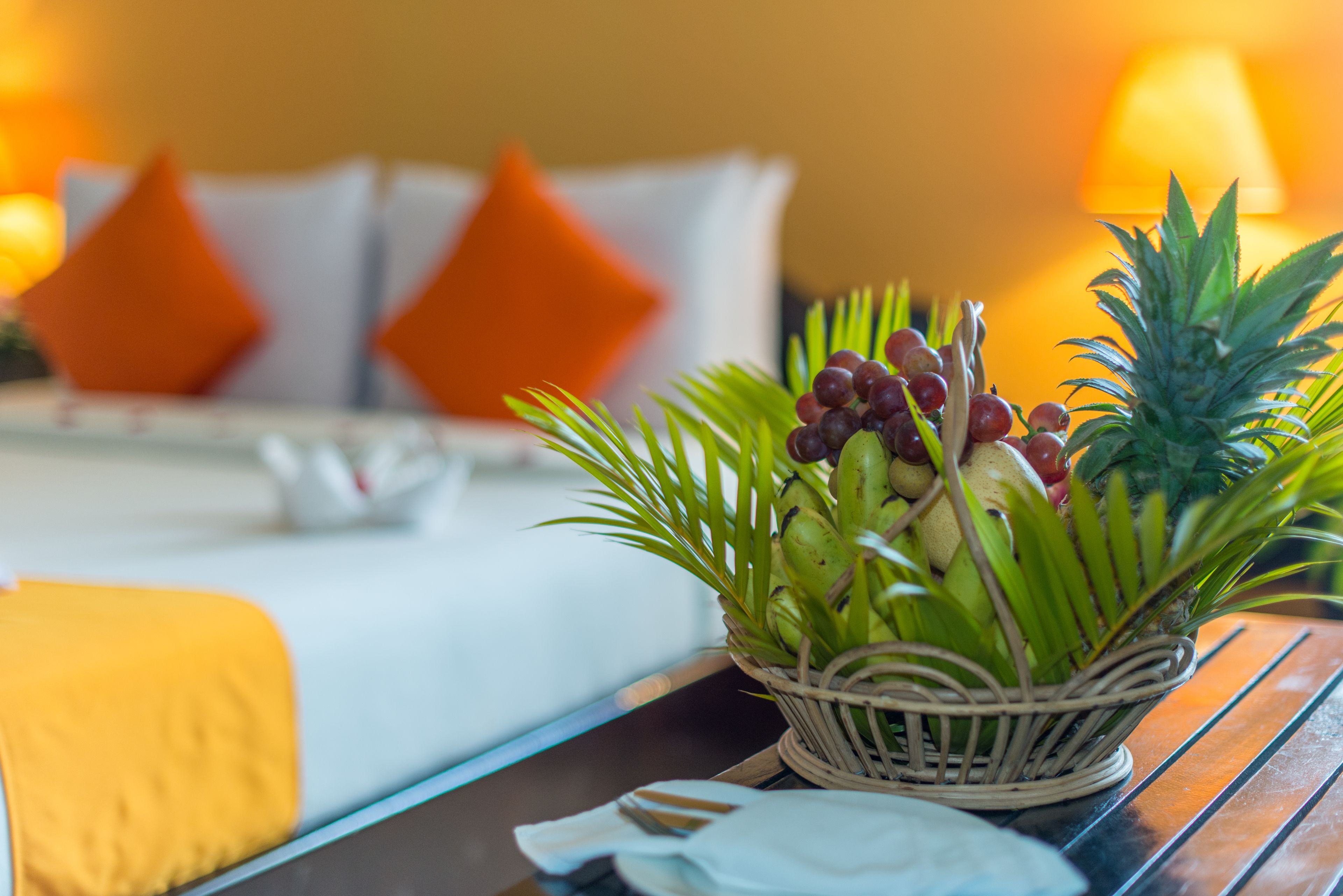 Una habitación de hotel con cama, almohadas, una cesta de frutas y una mesita de noche.