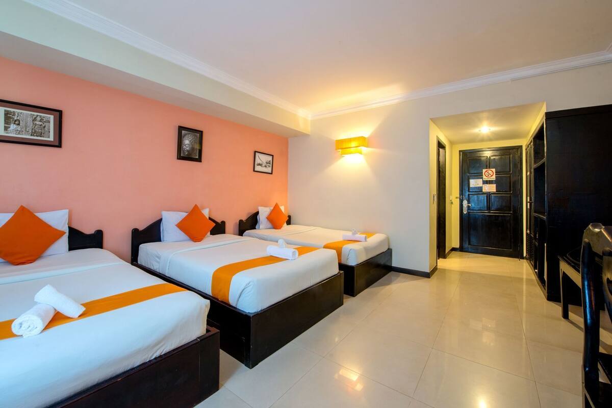 Ein Hotelzimmer mit drei Betten, jedes mit weißer Bettwäsche und orangen Akzenten, einem Flur mit einer Tür und einem Fernseher auf der rechten Seite.
