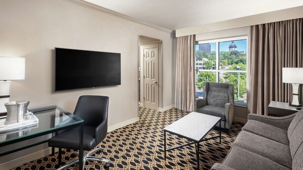 A hotel room with a large TV, a glass desk, a sofa, and a view of a building through the window.