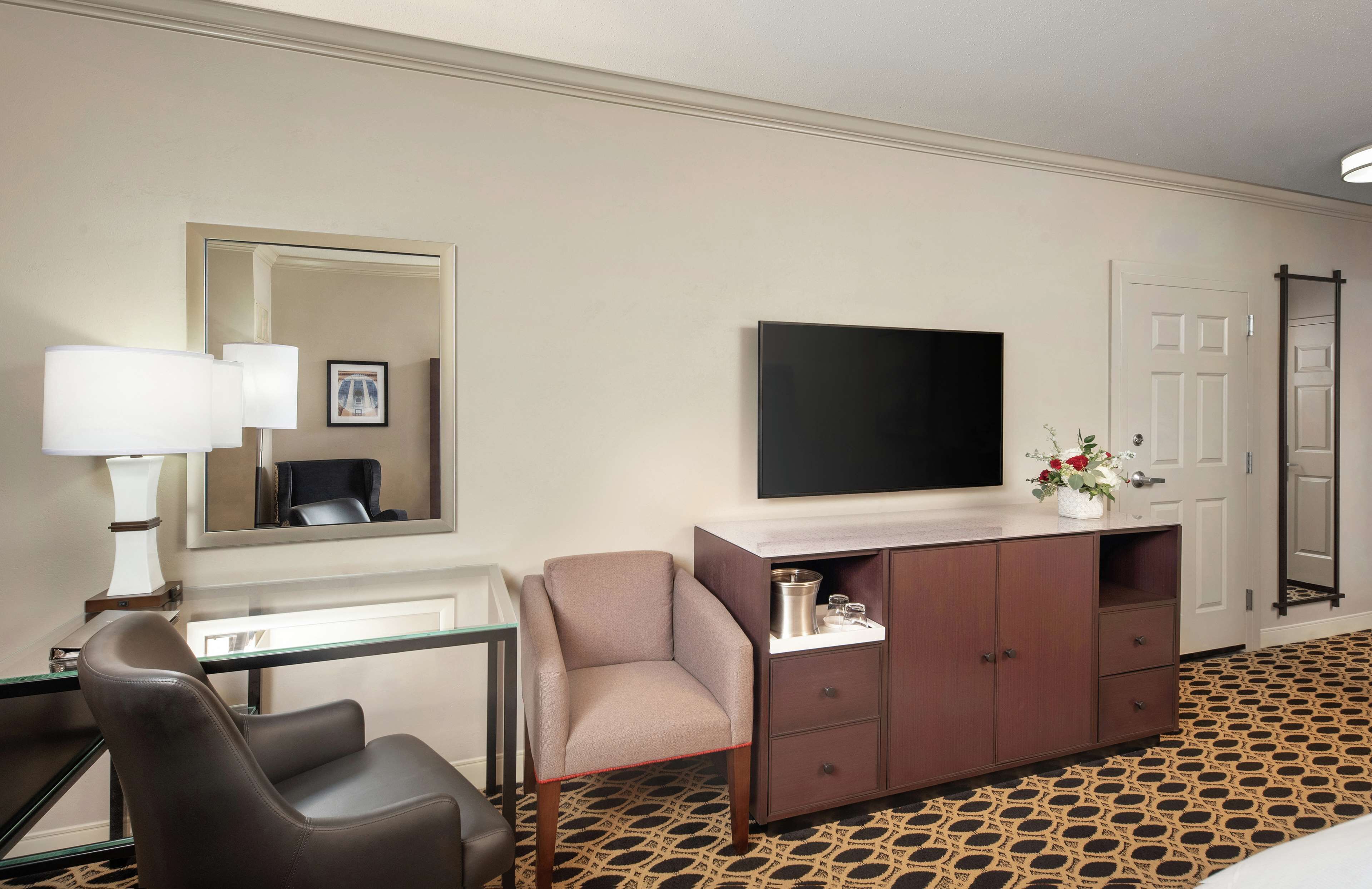 A hotel room with a bed, a desk with a lamp, a chair, a TV, a cabinet, and a patterned carpet.