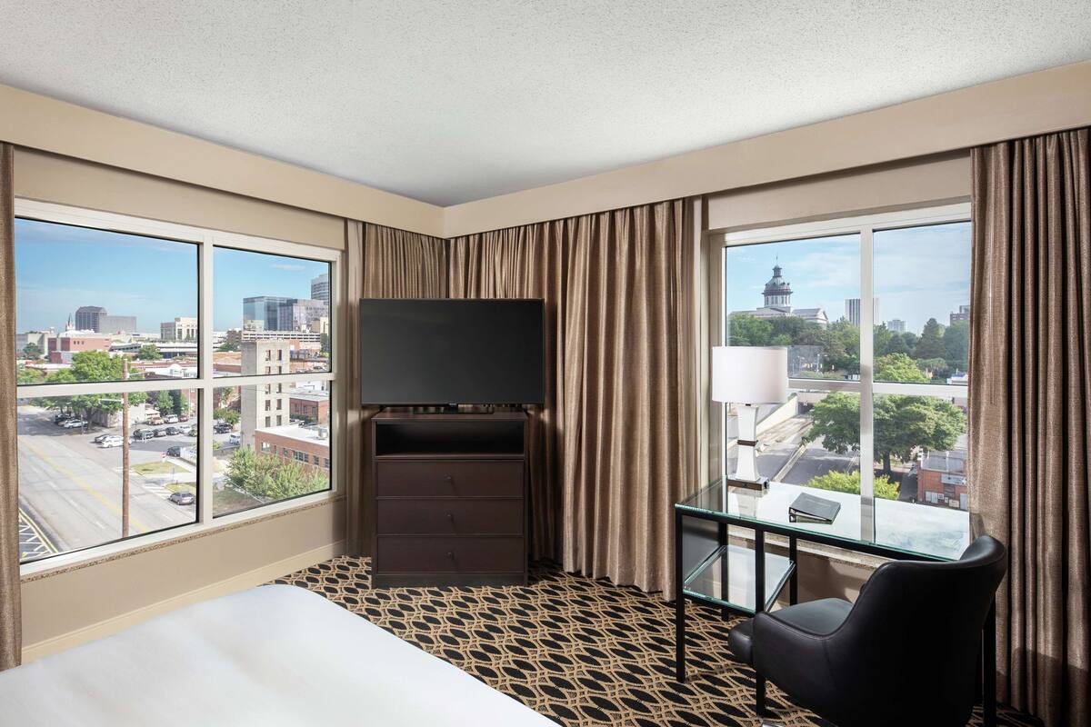 Camera d'albergo con un letto, una TV, una scrivania con una sedia e ampie finestre con vista sulla città.
