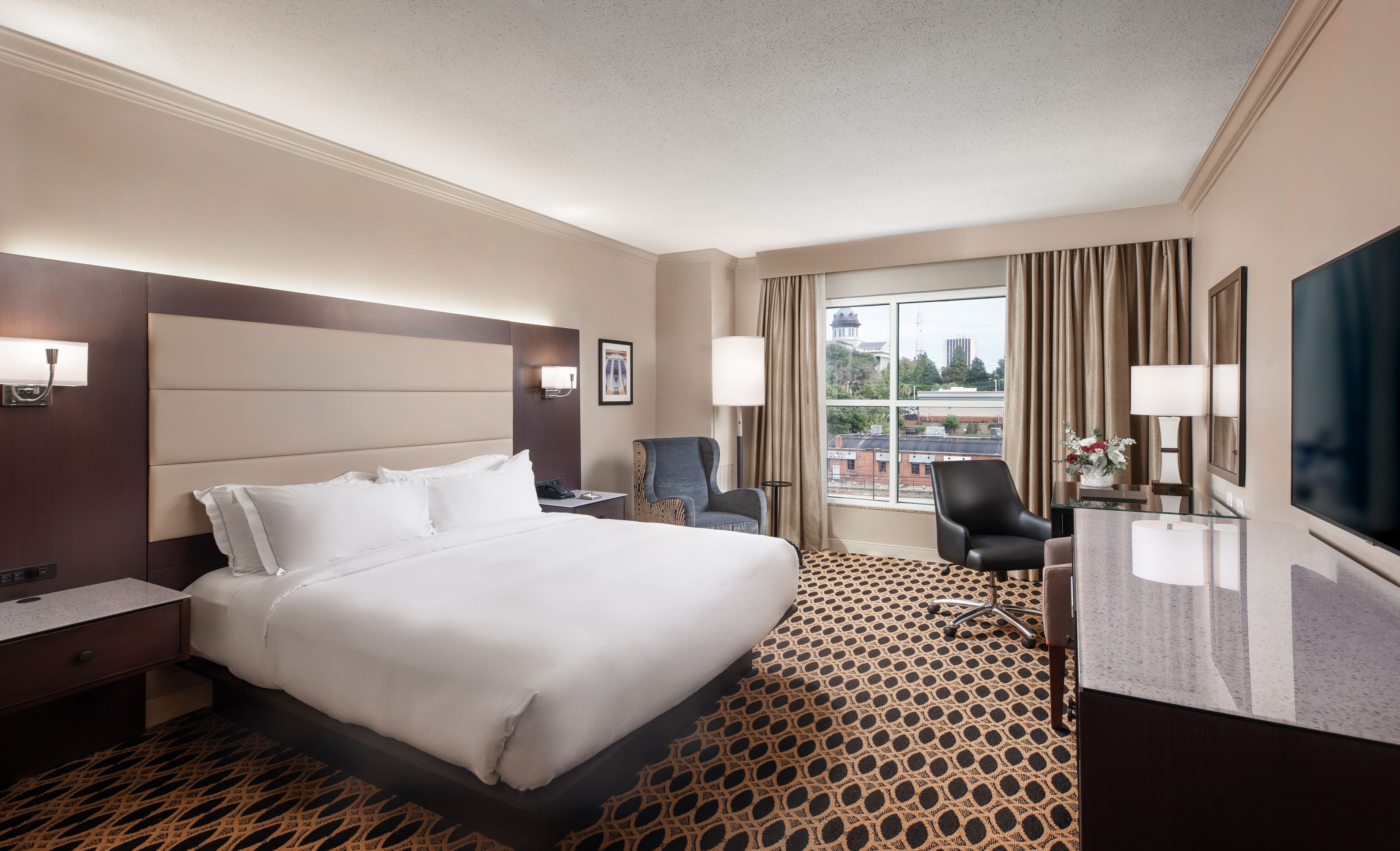 Une chambre d’hôtel avec un grand lit, un bureau, une chaise, une télévision et une fenêtre avec des rideaux.