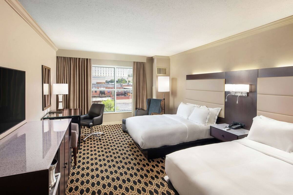 A hotel room with two beds, a desk with a TV, a chair, and a window with curtains.